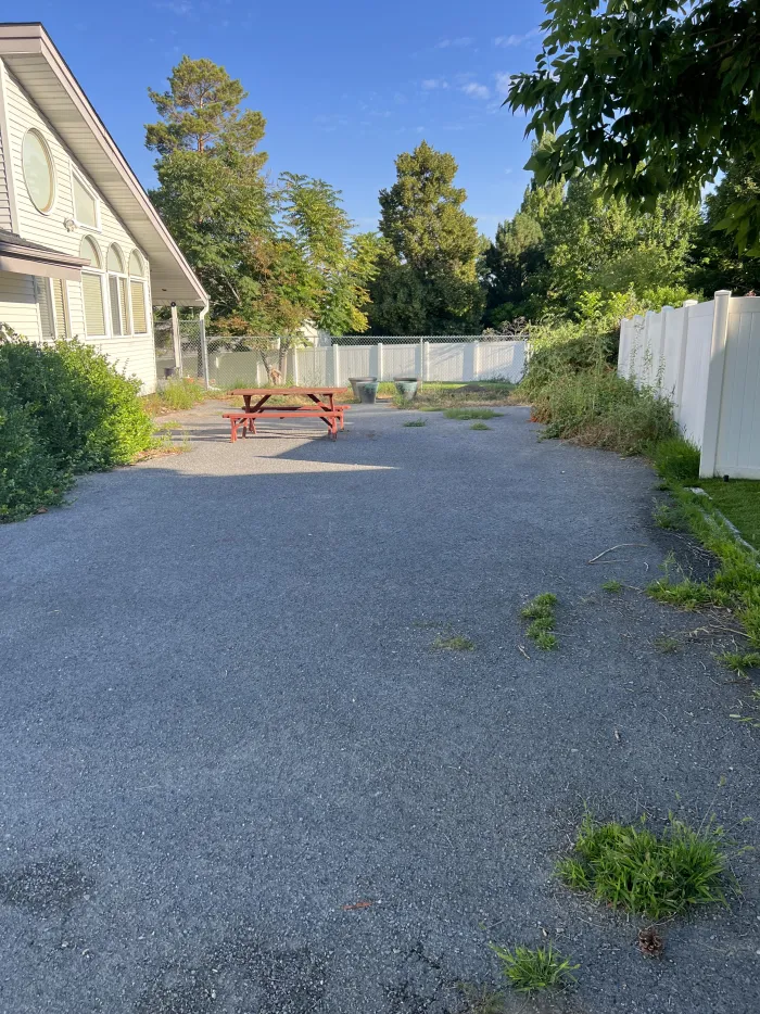 An old asphalt driveway with weeds, running along a white vinyl fence to a picnic table.