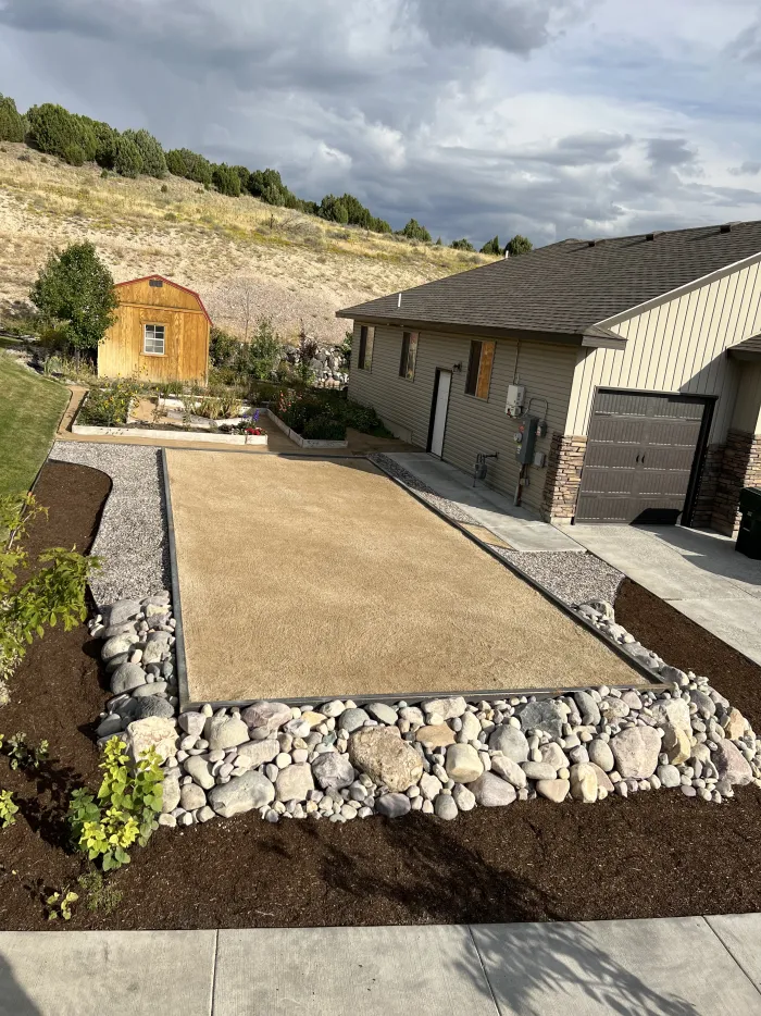 Bocce ball court of fine gravel bordered by river rock and mulch beside a house.