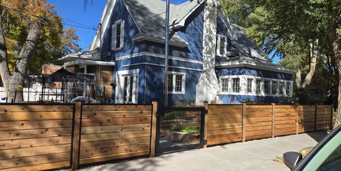 Horizontal cedar fence with a gate along the sidewalk in front of a blue Victorian house.