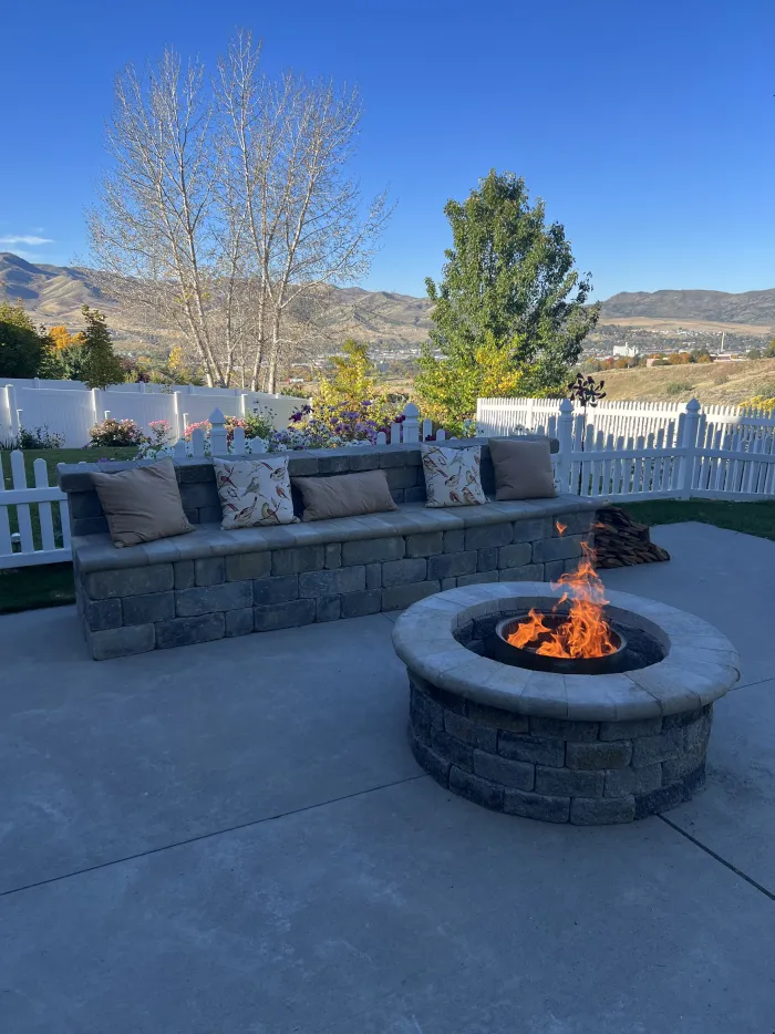 Round block fire pit with a fire burning, in front of a curved seat wall with cushions.