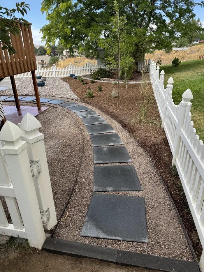 Walkway of large square gray pavers set in gravel, curving along a white picket fence.