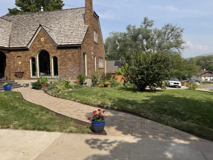 Curving paver walkway through a green lawn to a brick Tudor house, with red geraniums in a blue pot.