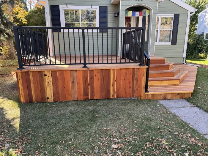 Redwood front deck with a black metal railing and steps, on a green house.