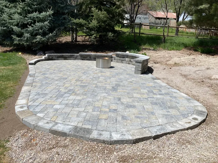 Curved paver patio edged in stone, with a block seat wall and fire pit, set in a large backyard.